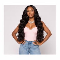 A woman modeling a long, loose wave wig in black, styled with a light pink sweetheart corset top and blue jeans against a light gray studio background, THE HAIR PUSHER PREMIUM LUXE HAIR.